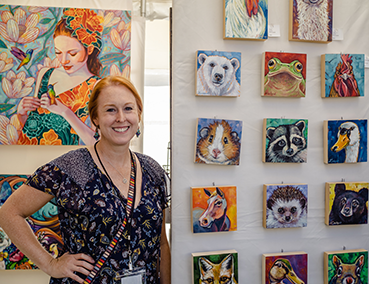 artist in her booth with brightly colored paintings of animals